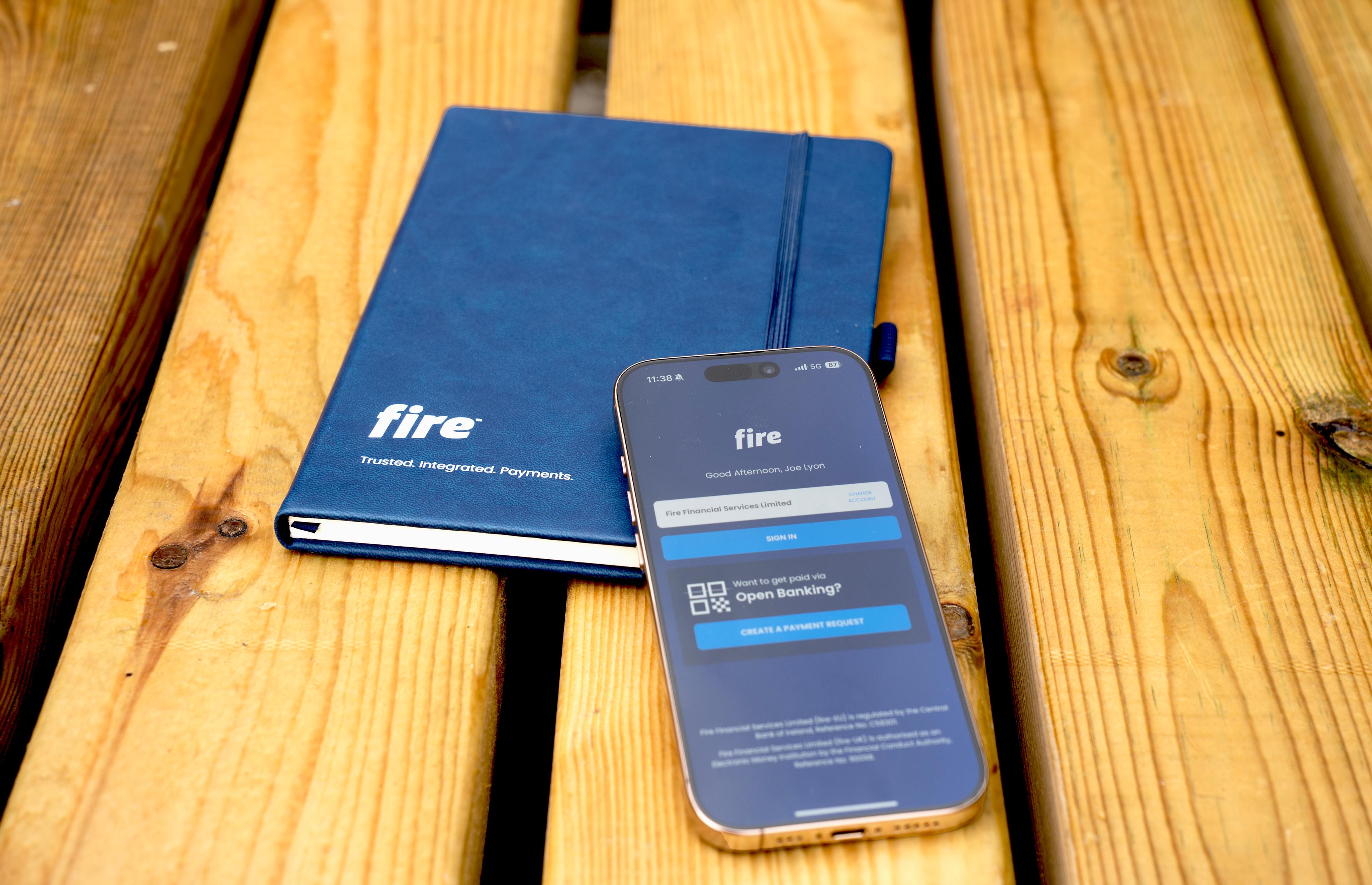 Phone displaying Fire’s login screen and an open banking payment request, placed over a notepad featuring Fire’s logo and the slogan “Trusted. Integrated. Payments.”